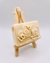 Load image into Gallery viewer, Handmade vegan soaps featuring a boy and girl sat on a bench with the wording Love Is... , in beautiful eco friendly packaging. crafted with natural French clays, activated charcoal and essential oils. a eco-friendly, zero waste gift that’s cruelty-free, skin-nourishing, the perfect gift for Valentines day or anniversary gift. small business UK