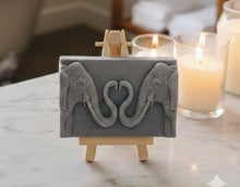 Load image into Gallery viewer, A rectangular handmade vegan soap featuring a detailed relief of two elephants facing each other with their trunks entwined to form a heart shape, displayed on a miniature wooden easel.