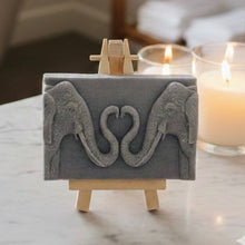 Load image into Gallery viewer, A rectangular handmade vegan soap featuring a detailed relief of two elephants facing each other with their trunks entwined to form a heart shape, displayed on a miniature wooden easel.