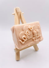 Load image into Gallery viewer, Handmade vegan soaps featuring a boy and girl sat on a bench with the wording Love Is... , in beautiful eco friendly packaging. crafted with natural French clays, activated charcoal and essential oils. a eco-friendly, zero waste gift that’s cruelty-free, skin-nourishing, the perfect gift for Valentines day or anniversary gift. small business UK