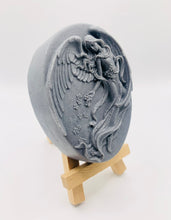 Load image into Gallery viewer, Handmade vegan Guardian Angel soap bar in a peach-toned French clay, featuring a detailed angel relief with flowing wings and flowers. Displayed on a small wooden easel. Natural, cruelty-free UK soap ideal for spiritual gifts, zero waste skincare, and sustainable bathroom décor. British-made artisan soap perfect for eco-conscious shoppers