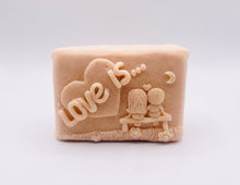 Load image into Gallery viewer, Handmade vegan soaps featuring a boy and girl sat on a bench with the wording Love Is... , in beautiful eco friendly packaging. crafted with natural French clays, activated charcoal and essential oils. a eco-friendly, zero waste gift that’s cruelty-free, skin-nourishing, the perfect gift for Valentines day or anniversary gift. small business UK