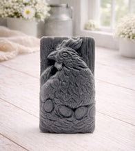 Load image into Gallery viewer, A rectangular handmade vegan soap in a natural colour, featuring a highly detailed relief carving of a broody hen chicken sitting on a nest of eggs, displayed upright on a light wooden surface.