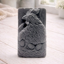 Load image into Gallery viewer, A rectangular handmade vegan soap in a natural colour, featuring a highly detailed relief carving of a broody hen chicken sitting on a nest of eggs, displayed upright on a light wooden surface.