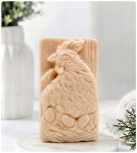 Load image into Gallery viewer, A rectangular handmade vegan soap in a natural colour, featuring a highly detailed relief carving of a broody hen chicken sitting on a nest of eggs, displayed upright on a light wooden surface.