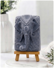 Load image into Gallery viewer, A rectangular handmade vegan soap in a speckled slate-grey finish, featuring a highly detailed relief of an elephant's head adorned with ornate, traditional patterns and a decorative floral background, displayed on a miniature wooden easel.