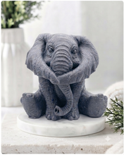 Load image into Gallery viewer, A highly detailed handmade vegan soap in a charcoal grey tone, sculpted in the shape of a sitting elephant with realistic wrinkled skin textures and large ears, displayed on a light wooden surface with white flowers in the background.