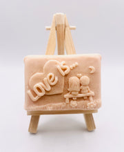 Load image into Gallery viewer, Handmade vegan soaps featuring a boy and girl sat on a bench with the wording Love Is... , in beautiful eco friendly packaging. crafted with natural French clays, activated charcoal and essential oils. a eco-friendly, zero waste gift that’s cruelty-free, skin-nourishing, the perfect gift for Valentines day or anniversary gift. small business UK
