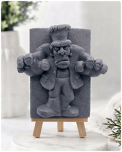 Load image into Gallery viewer, A rectangular handmade vegan soap featuring a high-relief carving of Frankenstein's monster in a classic pose with arms outstretched and a grumpy expression, displayed on a miniature wooden easel.