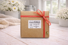 Load image into Gallery viewer, Gift box with a red ribbon on a wooden surface with blurred flowers in the background