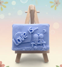Load image into Gallery viewer, Handmade vegan soaps featuring a boy and girl sat on a bench with the wording Love Is... , in beautiful eco friendly packaging. crafted with natural French clays, activated charcoal and essential oils. a eco-friendly, zero waste gift that’s cruelty-free, skin-nourishing, the perfect gift for Valentines day or anniversary gift. small business UK