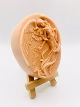 Load image into Gallery viewer, Handmade vegan Guardian Angel soap bar in a peach-toned French clay, featuring a detailed angel relief with flowing wings and flowers. Displayed on a small wooden easel. Natural, cruelty-free UK soap ideal for spiritual gifts, zero waste skincare, and sustainable bathroom décor. British-made artisan soap perfect for eco-conscious shoppers