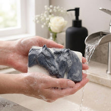 Load image into Gallery viewer, A rectangular handmade vegan soap featuring a detailed relief of two horse heads leaning affectionately towards each other, finished in a slate-grey tone against a white soap base and displayed on a miniature wooden easel.