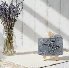 Load image into Gallery viewer, A spooky handmade vegan soap featuring a 3D relief of a skeleton relaxing against "RIP" tombstones, displayed on a wooden easel against a whimsical farmhouse decor background.