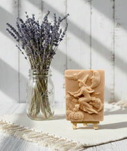 Load image into Gallery viewer, A beautiful handmade vegan soap featuring a detailed image of a witch flying on a broomstick past a crescent moon and owls, displayed on a wooden easel against a whimsical farmhouse decor background.