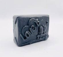 Load image into Gallery viewer, Handmade vegan soaps featuring a boy and girl sat on a bench with the wording Love Is... , in beautiful eco friendly packaging. crafted with natural French clays, activated charcoal and essential oils. a eco-friendly, zero waste gift that’s cruelty-free, skin-nourishing, the perfect gift for Valentines day or anniversary gift. small business UK