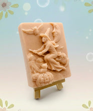 Load image into Gallery viewer, A beautiful handmade vegan soap featuring a detailed image of a witch flying on a broomstick past a crescent moon and owls, displayed on a wooden easel against a whimsical farmhouse decor background.