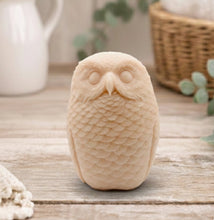 Load image into Gallery viewer, A detailed handmade vegan soap in the shape of an owl with intricate feather textures, displayed on a light wooden table with white flowers in the background.