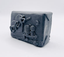 Load image into Gallery viewer, Handmade vegan soaps featuring a boy and girl sat on a bench with the wording Love Is... , in beautiful eco friendly packaging. crafted with natural French clays, activated charcoal and essential oils. a eco-friendly, zero waste gift that’s cruelty-free, skin-nourishing, the perfect gift for Valentines day or anniversary gift. small business UK