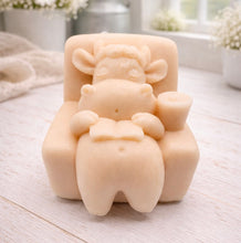 Load image into Gallery viewer, A handmade vegan soap sculpted to show a whimsical "lazy cow" character relaxing in an armchair, reading a book and holding a drink, displayed on a light wooden surface.