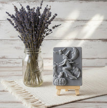 Load image into Gallery viewer, A beautiful handmade vegan soap featuring a detailed image of a witch flying on a broomstick past a crescent moon and owls, displayed on a wooden easel against a whimsical farmhouse decor background.