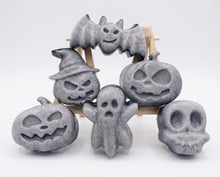 Load image into Gallery viewer, A collection of six handmade vegan Halloween soaps in a mottled grey and black charcoal finish, featuring a smiling bat, three different carved pumpkins (one wearing a witch's hat), a ghost, and a skull, arranged on and around a small wooden easel.