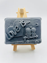 Load image into Gallery viewer, Handmade vegan soaps featuring a boy and girl sat on a bench with the wording Love Is... , in beautiful eco friendly packaging. crafted with natural French clays, activated charcoal and essential oils. a eco-friendly, zero waste gift that’s cruelty-free, skin-nourishing, the perfect gift for Valentines day or anniversary gift. small business UK