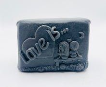 Load image into Gallery viewer, Handmade vegan soaps featuring a boy and girl sat on a bench with the wording Love Is... , in beautiful eco friendly packaging. crafted with natural French clays, activated charcoal and essential oils. a eco-friendly, zero waste gift that’s cruelty-free, skin-nourishing, the perfect gift for Valentines day or anniversary gift. small business UK