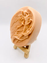 Load image into Gallery viewer, Handmade vegan Guardian Angel soap bar in a peach-toned French clay, featuring a detailed angel relief with flowing wings and flowers. Displayed on a small wooden easel. Natural, cruelty-free UK soap ideal for spiritual gifts, zero waste skincare, and sustainable bathroom décor. British-made artisan soap perfect for eco-conscious shoppers