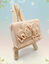 Load image into Gallery viewer, Handmade vegan soaps featuring a boy and girl sat on a bench with the wording Love Is... , in beautiful eco friendly packaging. crafted with natural French clays, activated charcoal and essential oils. a eco-friendly, zero waste gift that’s cruelty-free, skin-nourishing, the perfect gift for Valentines day or anniversary gift. small business UK