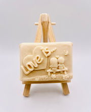 Load image into Gallery viewer, Handmade vegan soaps featuring a boy and girl sat on a bench with the wording Love Is... , in beautiful eco friendly packaging. crafted with natural French clays, activated charcoal and essential oils. a eco-friendly, zero waste gift that’s cruelty-free, skin-nourishing, the perfect gift for Valentines day or anniversary gift. small business UK