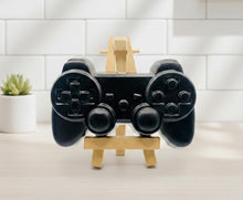 Load image into Gallery viewer, A 3D handmade vegan soap sculpted in the shape of a classic video game controller, finished in a deep charcoal black with realistic buttons and joysticks, displayed on a miniature wooden easel in a bright farmhouse setting.