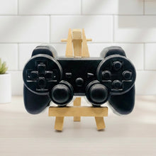 Load image into Gallery viewer, A 3D handmade vegan soap sculpted in the shape of a classic video game controller, finished in a deep charcoal black with realistic buttons and joysticks, displayed on a miniature wooden easel in a bright farmhouse setting.