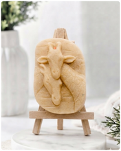 Load image into Gallery viewer, A rectangular handmade vegan soap in a natural cream tone, featuring a detailed relief of a mother and baby giraffe with their necks intertwined, displayed on a miniature wooden easel against a white wood-panelled background.