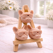 Load image into Gallery viewer, A set of three handmade vegan soaps in the shape of French Bulldog heads wearing sunglasses, displayed on a wooden easel in a bright, modern bathroom setting.