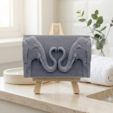 Load image into Gallery viewer, A rectangular handmade vegan soap featuring a detailed relief of two elephants facing each other with their trunks entwined to form a heart shape, displayed on a miniature wooden easel.