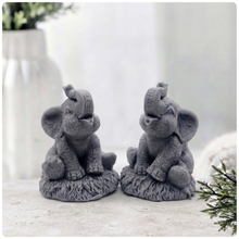 Load image into Gallery viewer, A charming gift set of two handmade vegan soaps, one sculpted as a sitting baby elephant with a trunk raised in a greeting and the other as a smaller standing calf, displayed on a light wooden surface in a bright farmhouse setting.