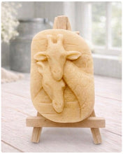 Load image into Gallery viewer, A rectangular handmade vegan soap in a natural tone, featuring a detailed relief of a mother and baby giraffe with their necks intertwined, displayed on a miniature wooden easel against a white wood-panelled background.