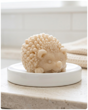 Load image into Gallery viewer, A detailed handmade vegan soap sculpted in the shape of a round, sitting hedgehog with intricate quill textures and a cute face, in a natural honey-brown tone, displayed on a light wooden surface.