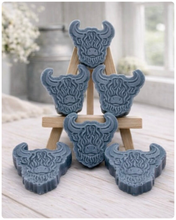 Load image into Gallery viewer, A collection of six handmade vegan soaps each intricately sculpted into the head of a shaggy Highland cow with long horns and a thick, textured coat, arranged in a pyramid on and around a small wooden easel.