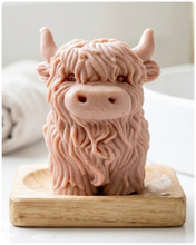 Load image into Gallery viewer, A detailed handmade vegan soap in a sculpted in the shape of a highland cow with characteristic long, wavy fur, horns, and a friendly face, displayed on a light wooden surface