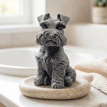 Load image into Gallery viewer, A detailed handmade vegan soap sculpted in the shape of a sitting Schnauzer dog with characteristic fluffy eyebrows and beard, in a natural colour, set against a bright window and white flowers.