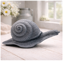 Load image into Gallery viewer, A highly detailed handmade vegan soap in a slate-grey tone, sculpted in the shape of a realistic snail with a prominent spiralled shell and a textured, leathery body, displayed on a light wooden surface.