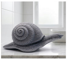 Load image into Gallery viewer, A highly detailed handmade vegan soap in a slate-grey tone, sculpted in the shape of a realistic snail with a prominent spiralled shell and a textured, leathery body, displayed on a light wooden surface.