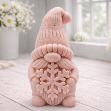 Load image into Gallery viewer, handmade vegan soap in the shape of a winter gnome sculpture holding a large snowflake, featuring a tall textured hat and a bushy moustache , displayed on a rustic white wooden surface with daisies and a milk churn in the background.