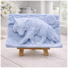 Load image into Gallery viewer, A rectangular handmade vegan soap, featuring a detailed relief carving of a wolf in mid-stride across a rugged landscape with a large full moon in the background, displayed on a miniature wooden easel.
