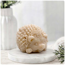 Load image into Gallery viewer, A detailed handmade vegan soap sculpted in the shape of a round, sitting hedgehog with intricate quill textures and a cute face, in a natural honey-brown tone, displayed on a light wooden surface.
