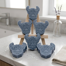 Load image into Gallery viewer, A collection of six handmade vegan soaps each intricately sculpted into the head of a shaggy Highland cow with long horns and a thick, textured coat, arranged in a pyramid on and around a small wooden easel.