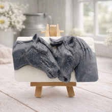 Load image into Gallery viewer, A rectangular handmade vegan soap featuring a detailed relief of two horse heads leaning affectionately towards each other, finished in a slate-grey tone against a white soap base and displayed on a miniature wooden easel.