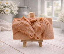 Load image into Gallery viewer, A rectangular handmade vegan soap featuring a detailed relief of two horse heads leaning affectionately towards each other, finished in soft natural tones and displayed on a miniature wooden easel.